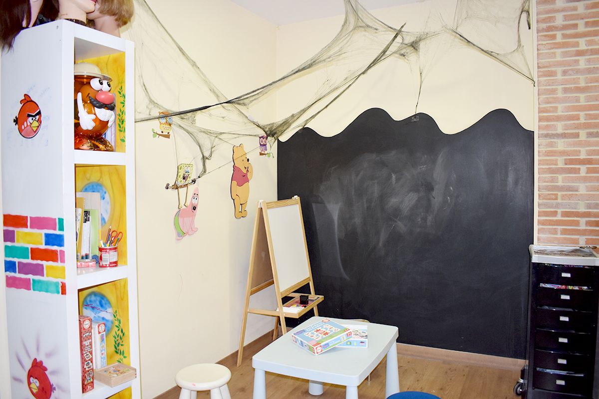 Zona infantil de la peluquería. Hay una estantería con juegos para niños, una mesa con dos taburetes y una pizarra blanca con patas y otra pizarra negra pegada en la pared para pintar.